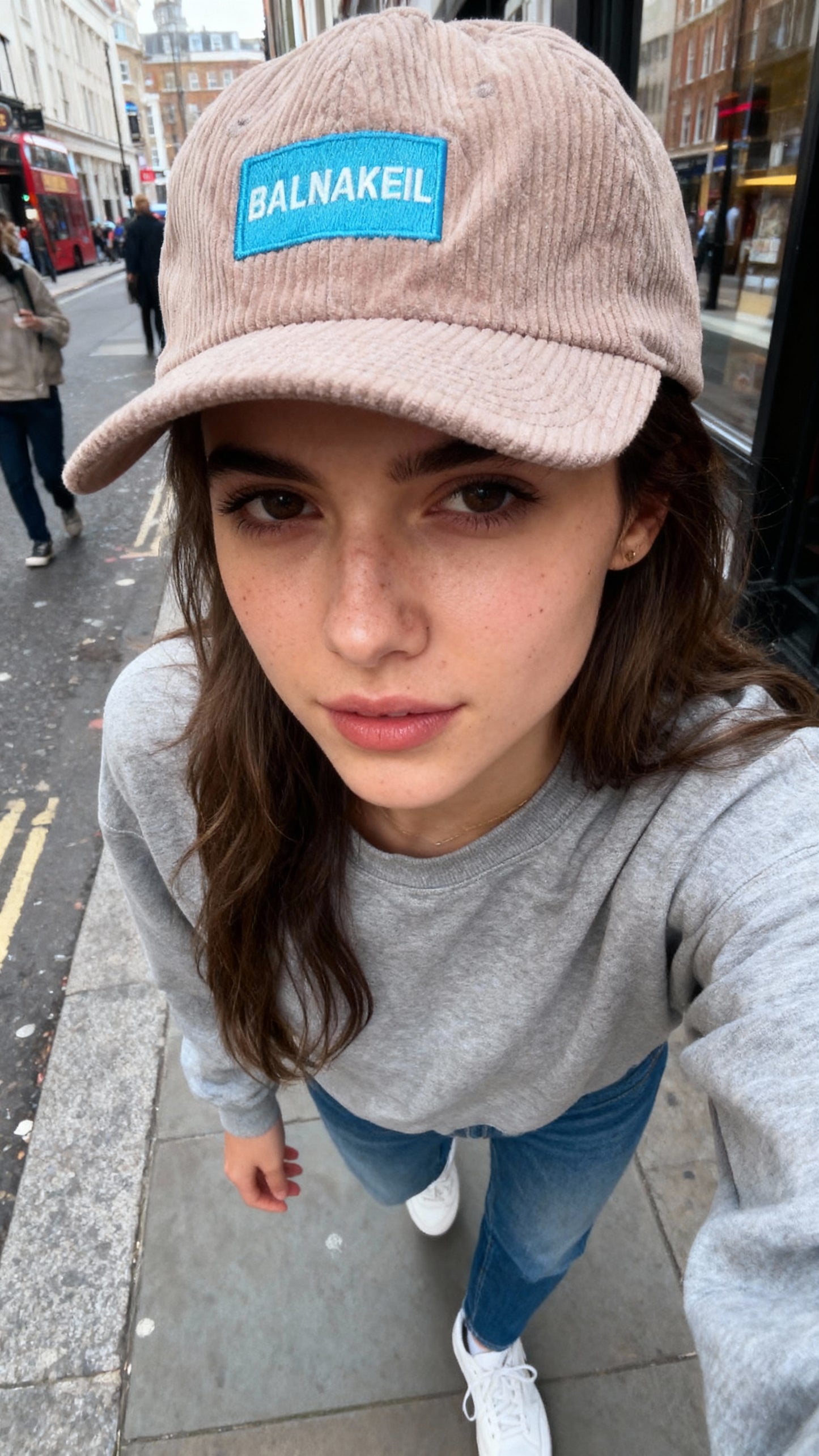 A women with brown hair taking a selfie with a pink Balnakeil cap on walking down a London street.