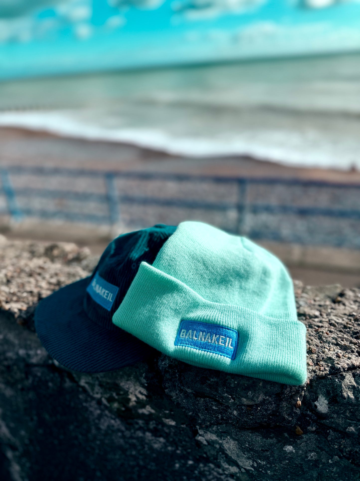 Two hats, one blue cord cap and one turquoise beanie, with a Balnakeil brand logo on a stone surface near water.