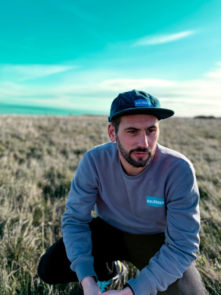 Man crouched wearing Balnakeil branded grey sweatshirt and teal cord cap with a blue sky.