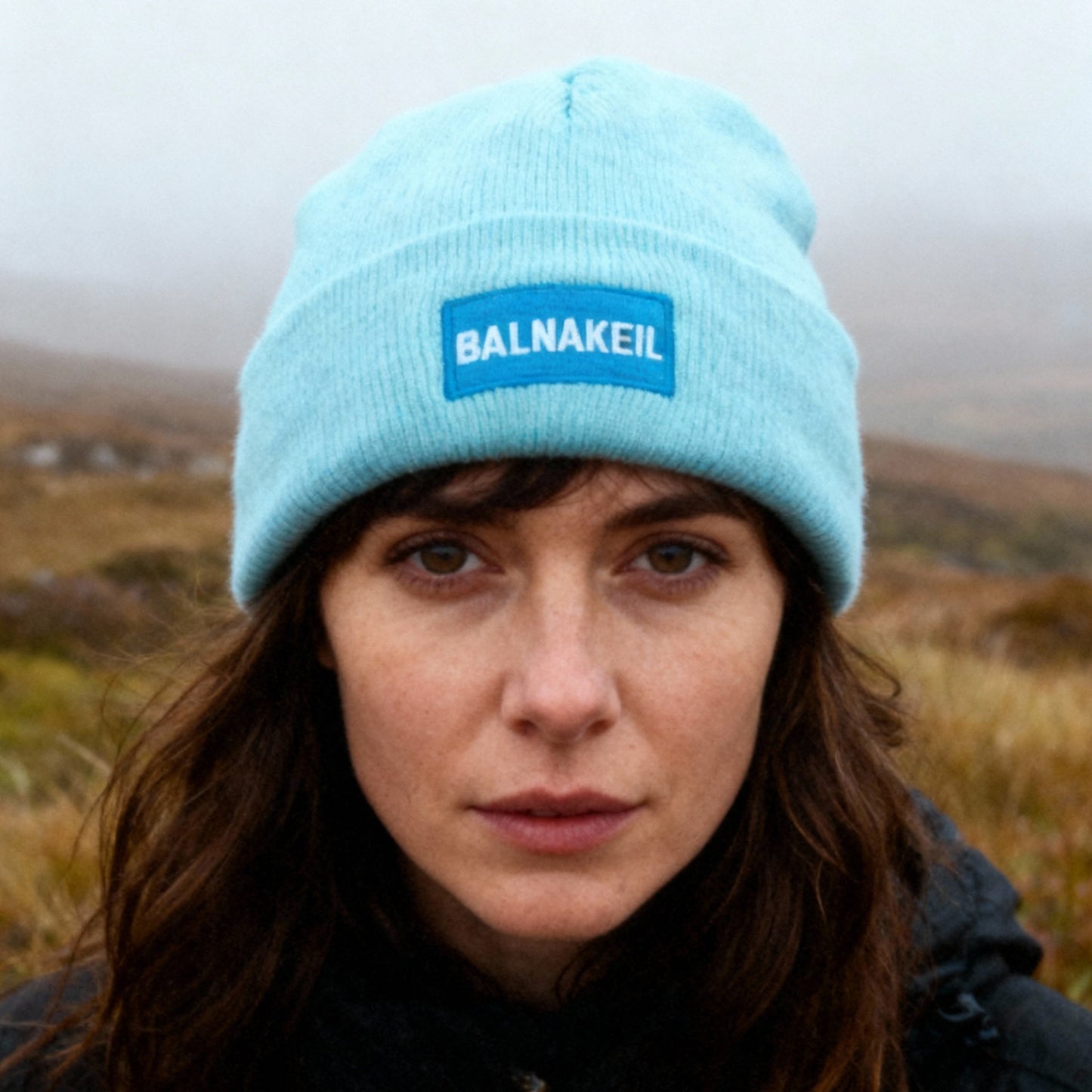 Person wearing a light blue beanie with 'Balnakeil' logo in a landscape setting