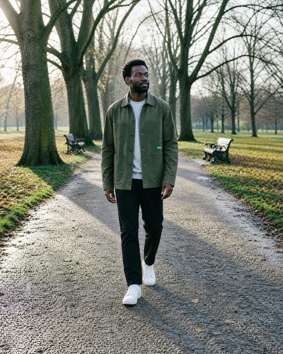 A man walking on a gravel path in a park, wearing an olive green overshirt over a white t-shirt, black trousers, and white sneakers.