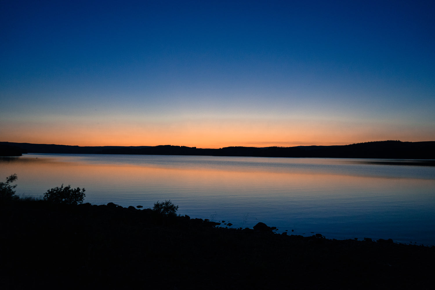 Sunset over a calm lake with silhouettes of trees and mountains.
