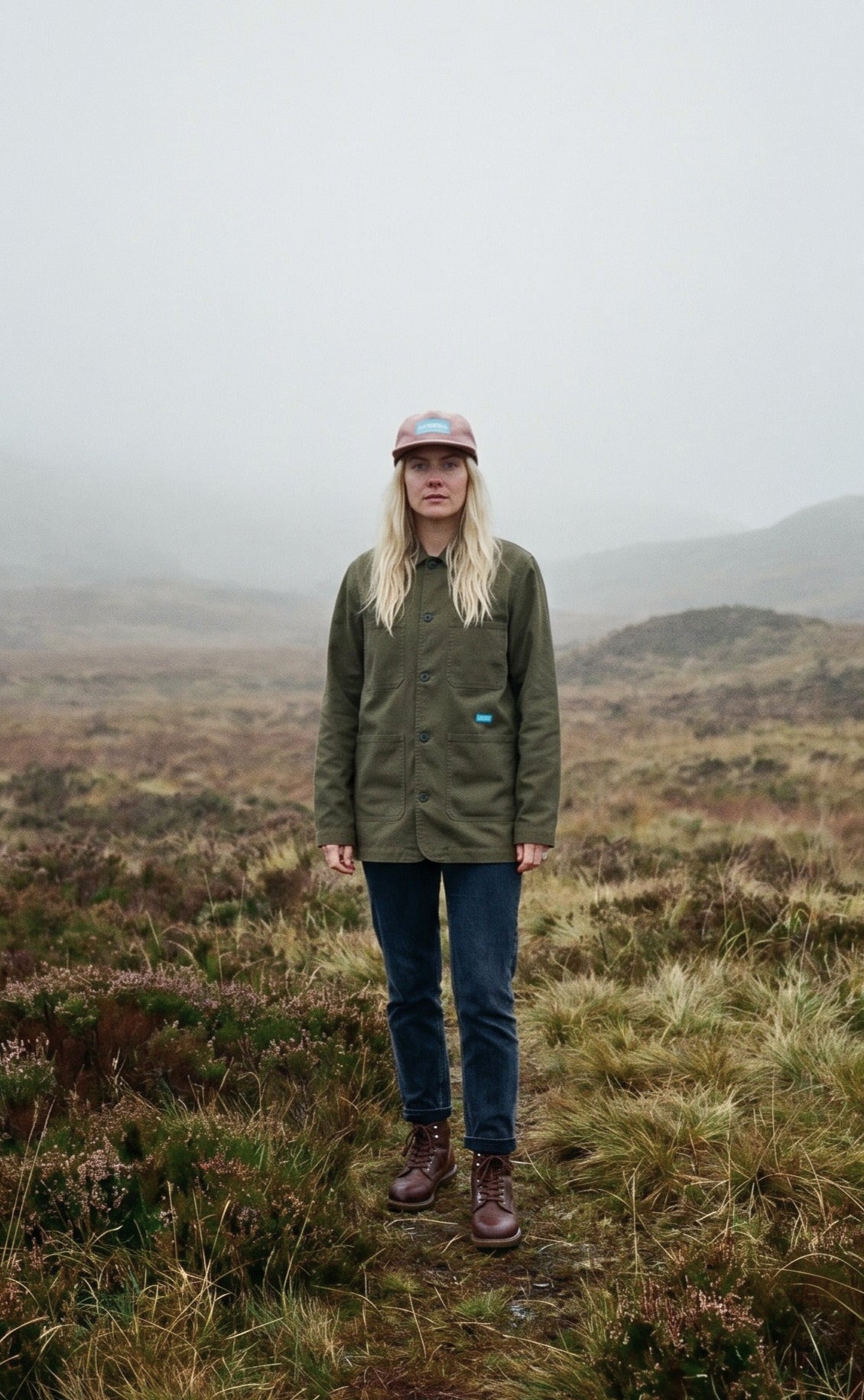 Person standing in a misty landscape wearing a green jacket and jeans.